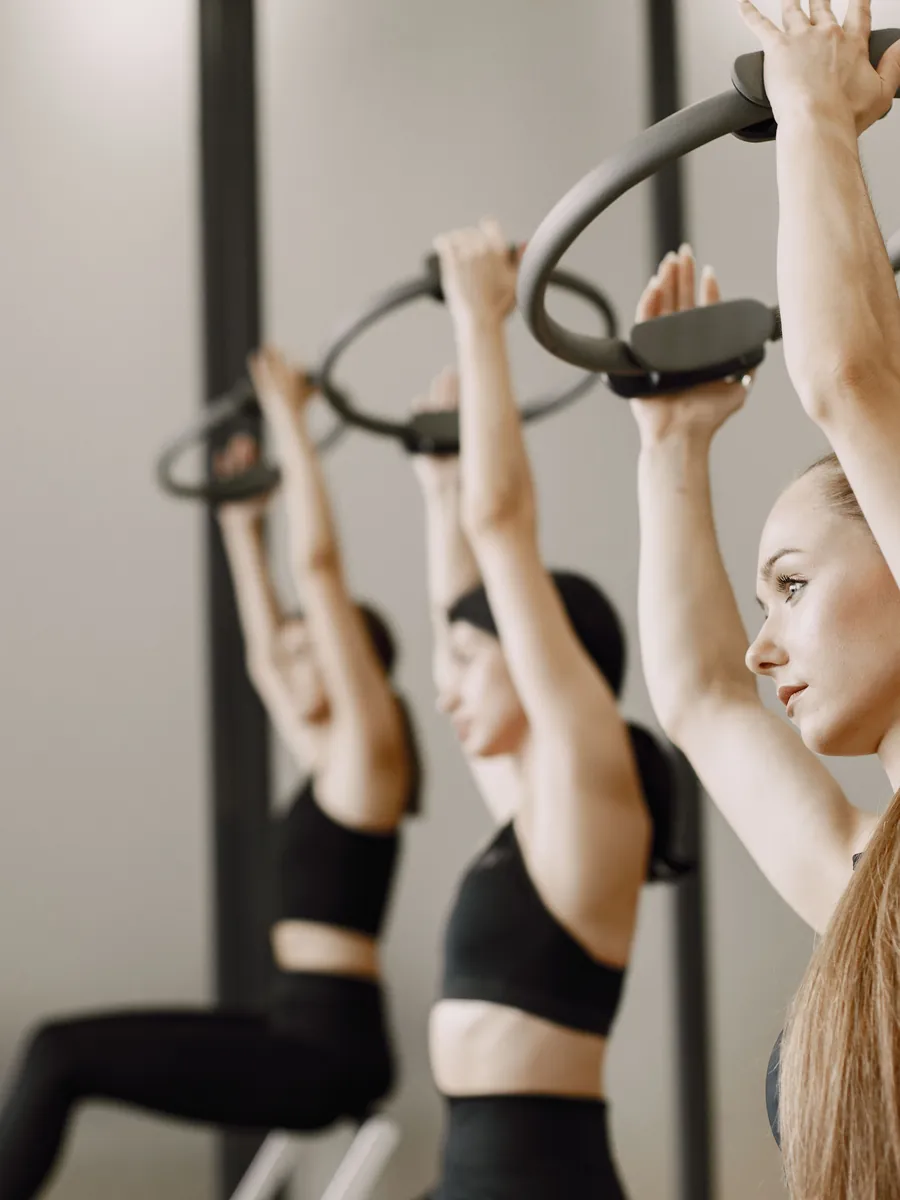 Alunas em pé sustentando o arco de pilates acima da cabeça.